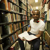 A man sits on a chair with a book on his lap in the stacks of a library.